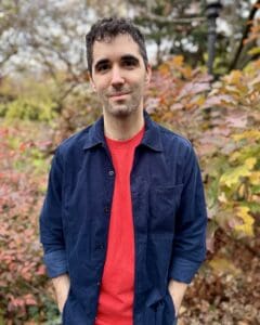 Smiling man in a navy overshirt and red shirt stands outdoors in a colorful autumn garden, surrounded by trees and foliage.