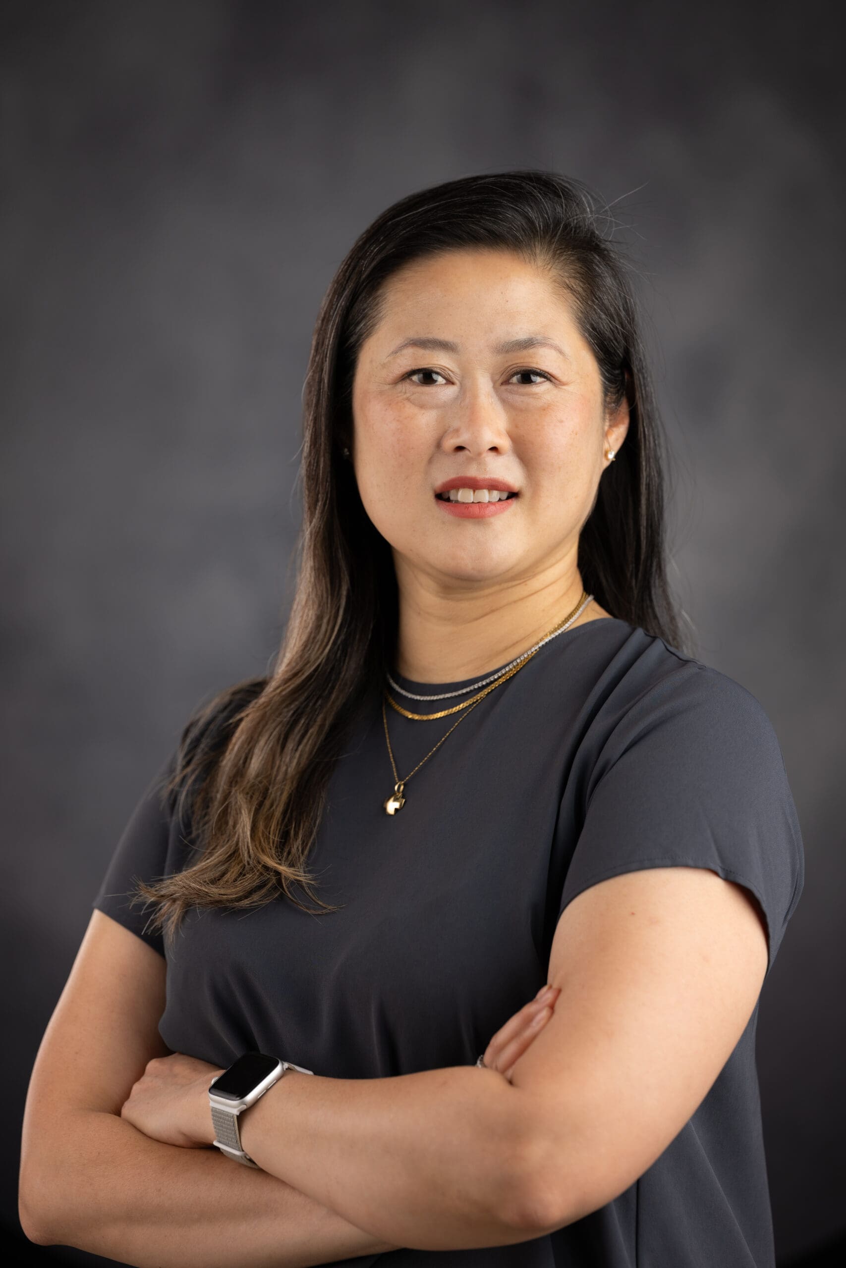 A person with long dark hair, wearing a dark gray top and layered necklaces, stands with arms crossed against a dark gray background.