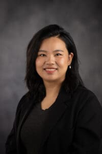 A smiling person with dark hair and a black blazer stands against a dark gray background.