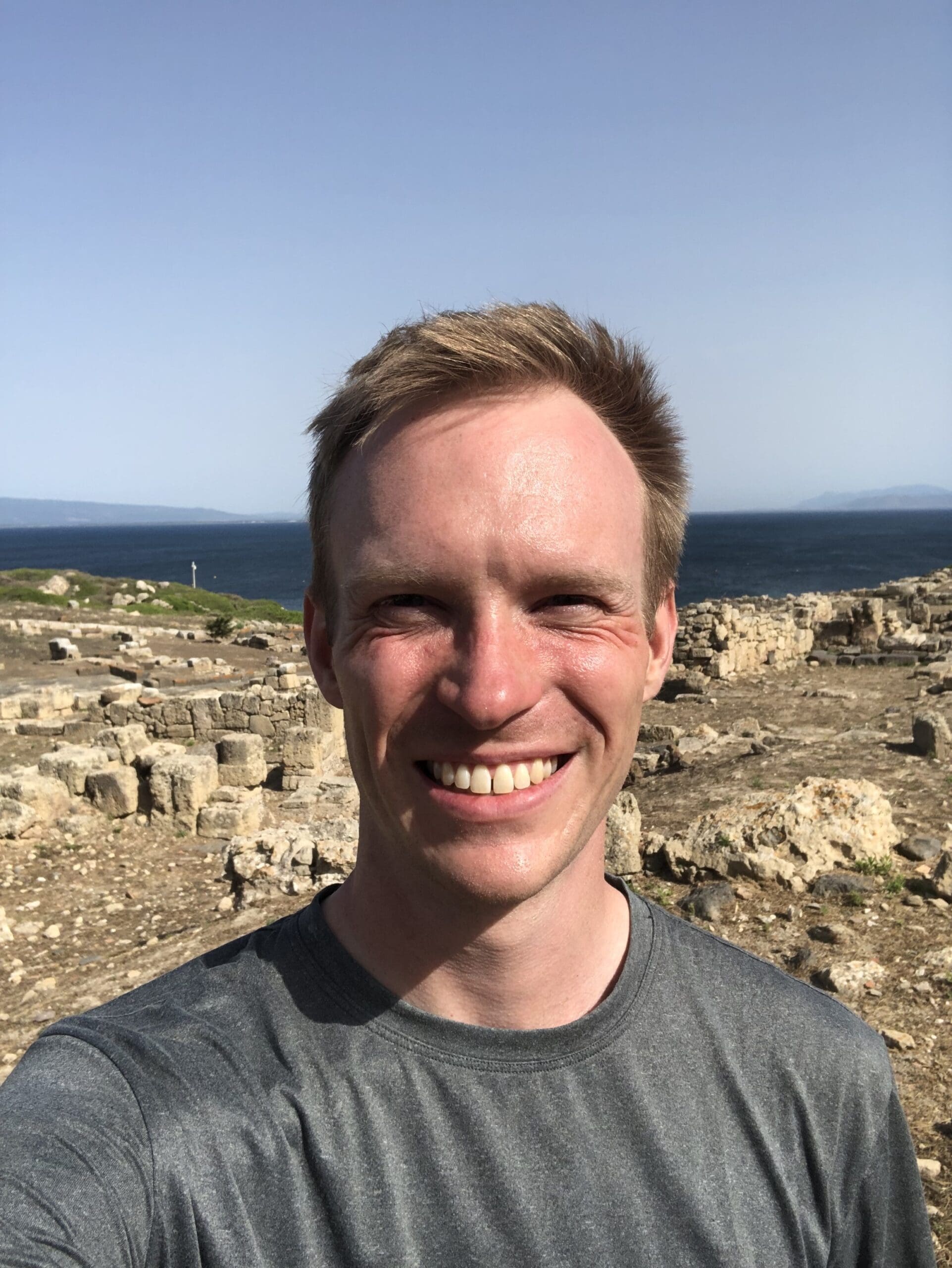 Outdoor portrait of a person in a dark T-shirt, standing near a rocky shoreline or coastal overlook with water in the distance.
