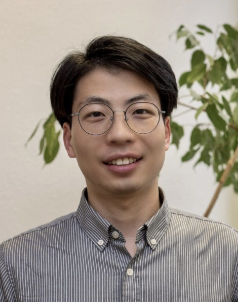 Zhiwei Wang A man looks into the camera smiling wearing a striped button down shirt