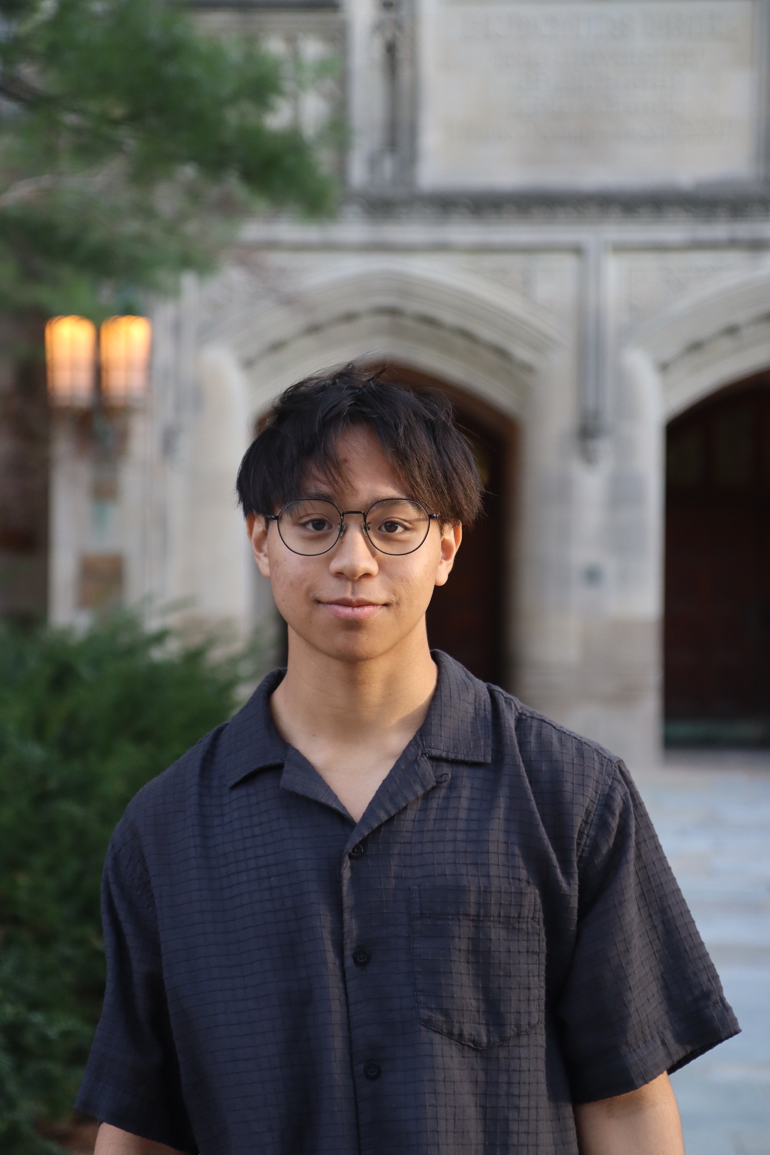 Young person wearing round glasses and a dark button-up shirt stands calmly before a historic stone archway, with warm lanterns glowing nearby.