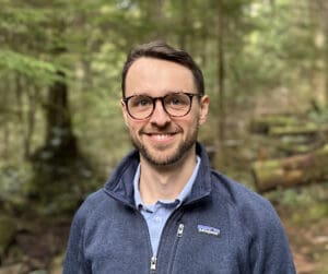 Man smiling at camera wearing glasses, forest behind him.