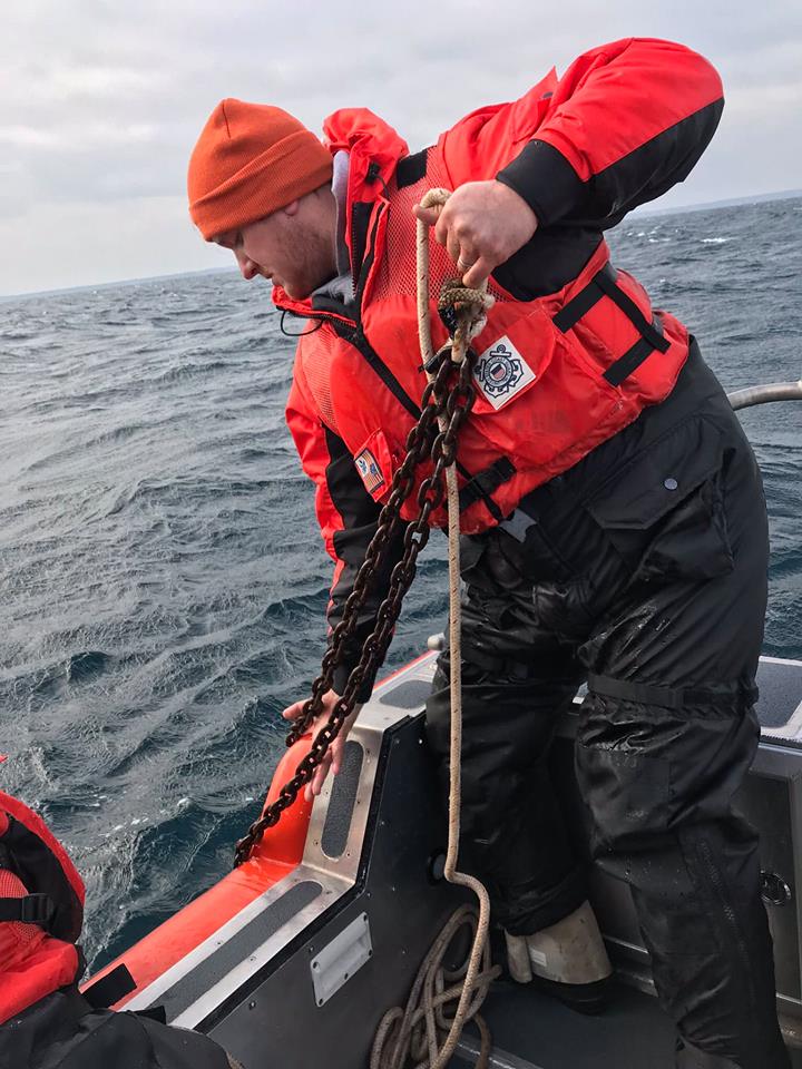 Person in a red life jacket hauls a heavy chain onto a boat on choppy water.