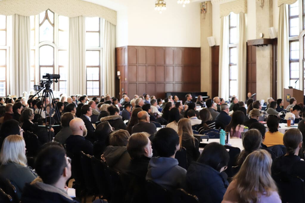 Wide view of a crowded auditorium with tall windows and a camera on a tripod recording the event