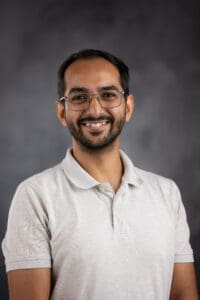 A person with glasses and a beard smiles warmly while wearing a light-colored polo shirt against a dark gray background.