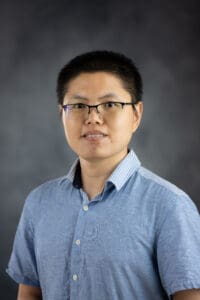 A person with short dark hair and glasses wears a light blue button-up shirt with a striped collar against a dark gray background.
