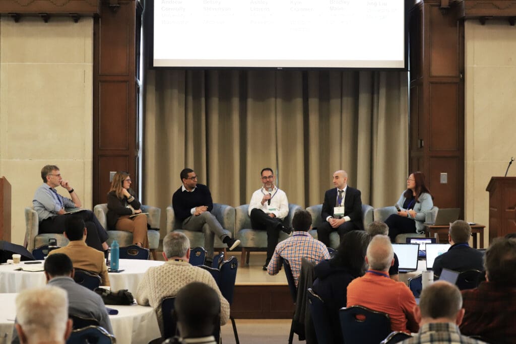 A panel of speakers sits on stage in front of a large audience during a conference session