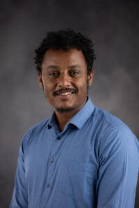 A person with curly dark hair and a mustache smiles warmly while wearing a blue button-up shirt against a dark gray background.