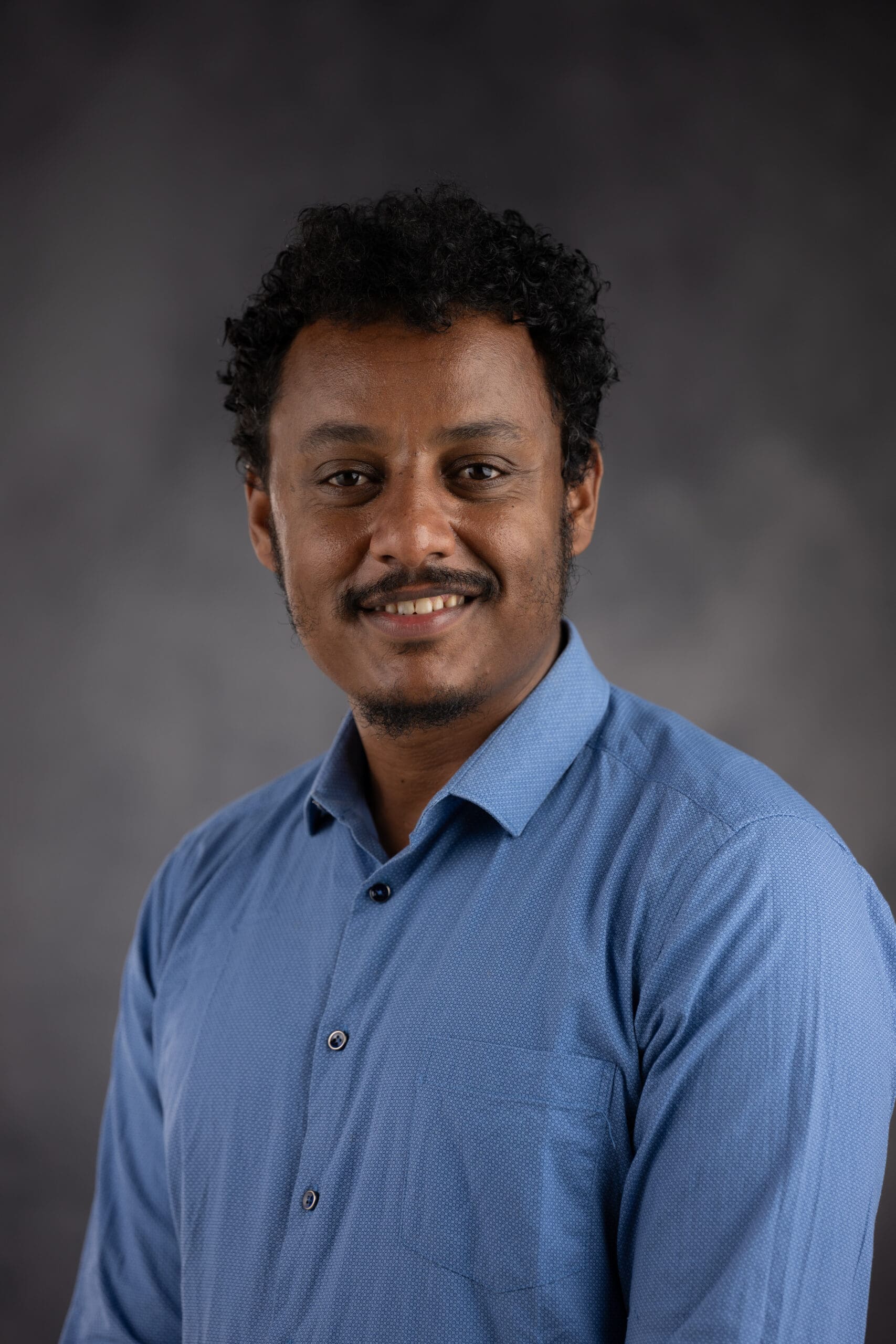 A person with curly dark hair and a mustache smiles warmly while wearing a blue button-up shirt against a dark gray background.
