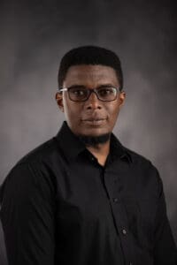 A person with short dark hair and glasses wears a black button-up shirt against a dark gray background.