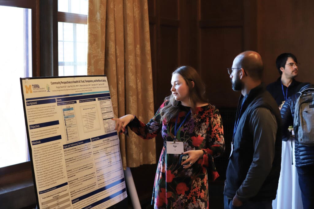A conference attendee gestures to a large research poster on an easel while speaking with two other attendees near a window.