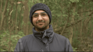 A headshot of a man standing in front of blurred green foliage