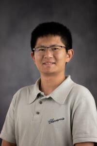 A person with short dark hair and glasses smiles while wearing a light-colored polo shirt with the word "Chavarria" embroidered on the chest, set against a plain gray background.