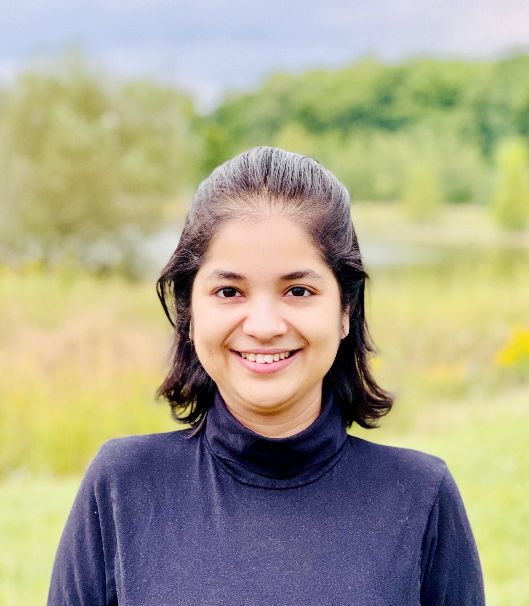 Outdoor portrait of a person in a blue turtleneck with trees and a field behind them