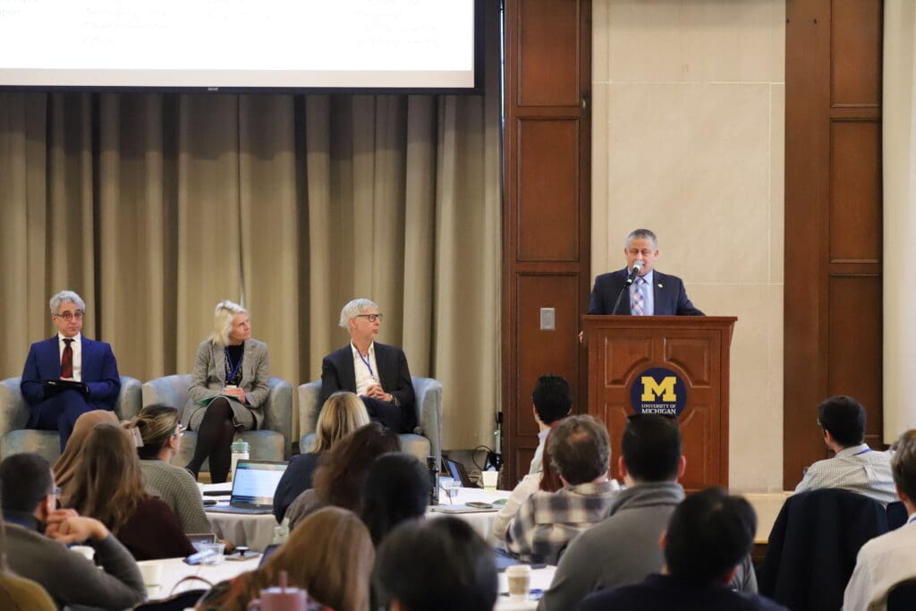 A speaker at a U‑M podium presents to the audience while three panelists sit to the side on stage