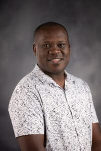 A person with short hair and a patterned shirt smiles gently against a dark, textured background.