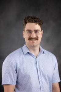 A person with curly hair, glasses, and a mustache smiles while wearing a light blue patterned short-sleeved shirt against a dark gray background.