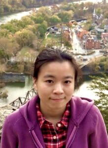 Portrait of a woman in a purple jacket at a scenic overlook, with an autumn river, wooded park, bridge, and town visible behind her.