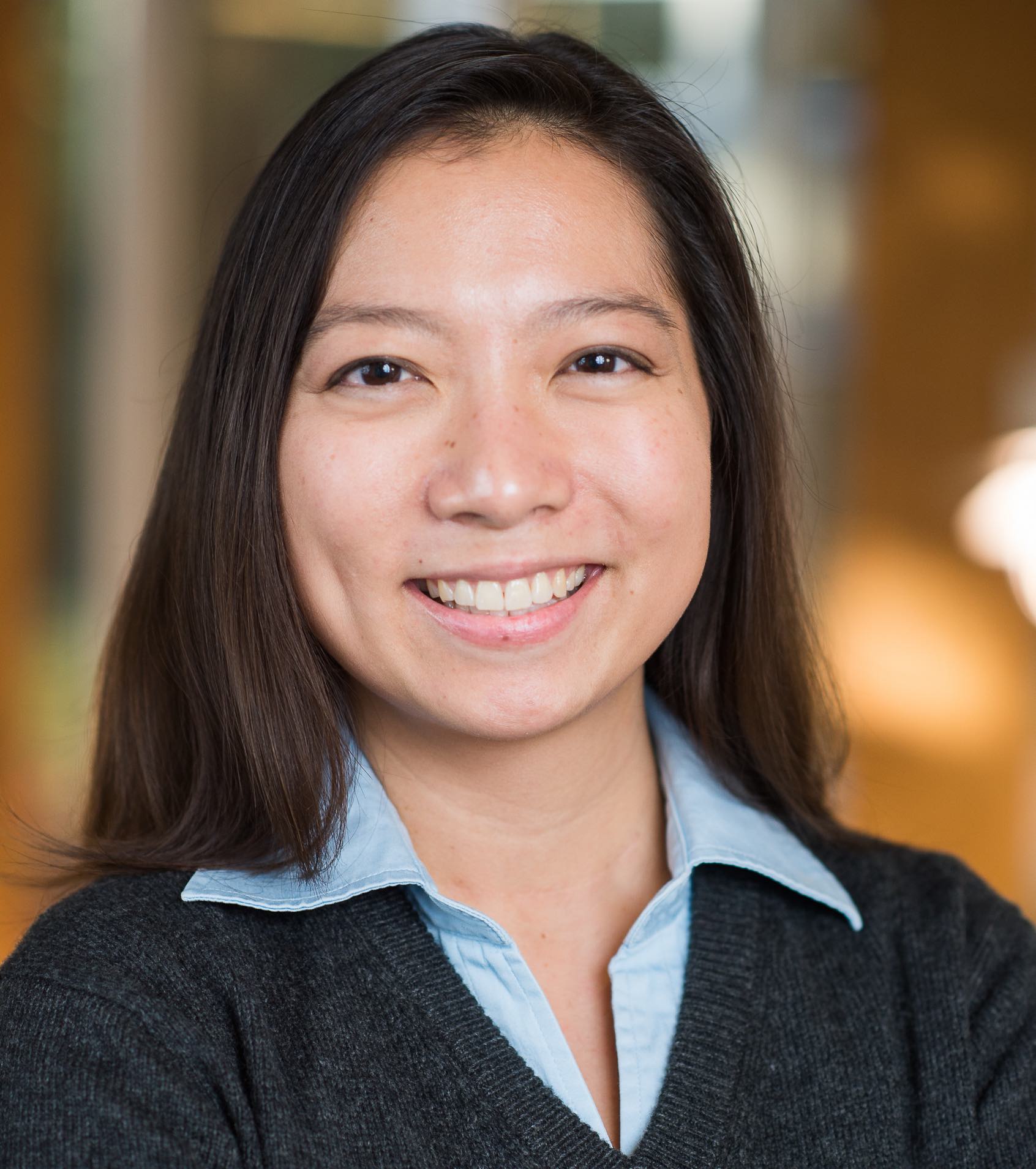 Smiling professional woman with long dark hair, wearing a light blue collared shirt and dark cardigan, posed against a softly blurred background.