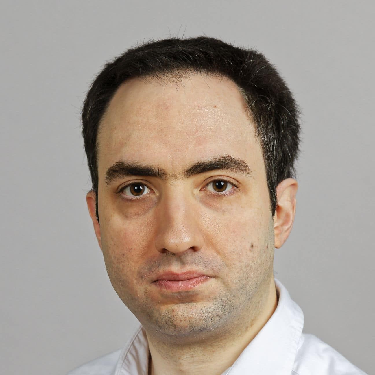 Head-and-shoulders portrait of a person with short dark hair, thick eyebrows, and a neutral expression, wearing a white collared shirt against a plain light gray background.