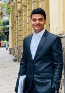 A person in a dark suit and white shirt stands on a city sidewalk beside a row of historic columns, smiling at the camera while holding a folder under one arm.