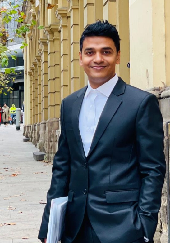 A person in a dark suit and white shirt stands on a city sidewalk beside a row of historic columns, smiling at the camera while holding a folder under one arm.