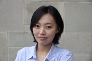 A person with short dark hair wears a light blue striped button-up shirt and a delicate necklace, smiling gently while standing in front of a light gray tiled wall.