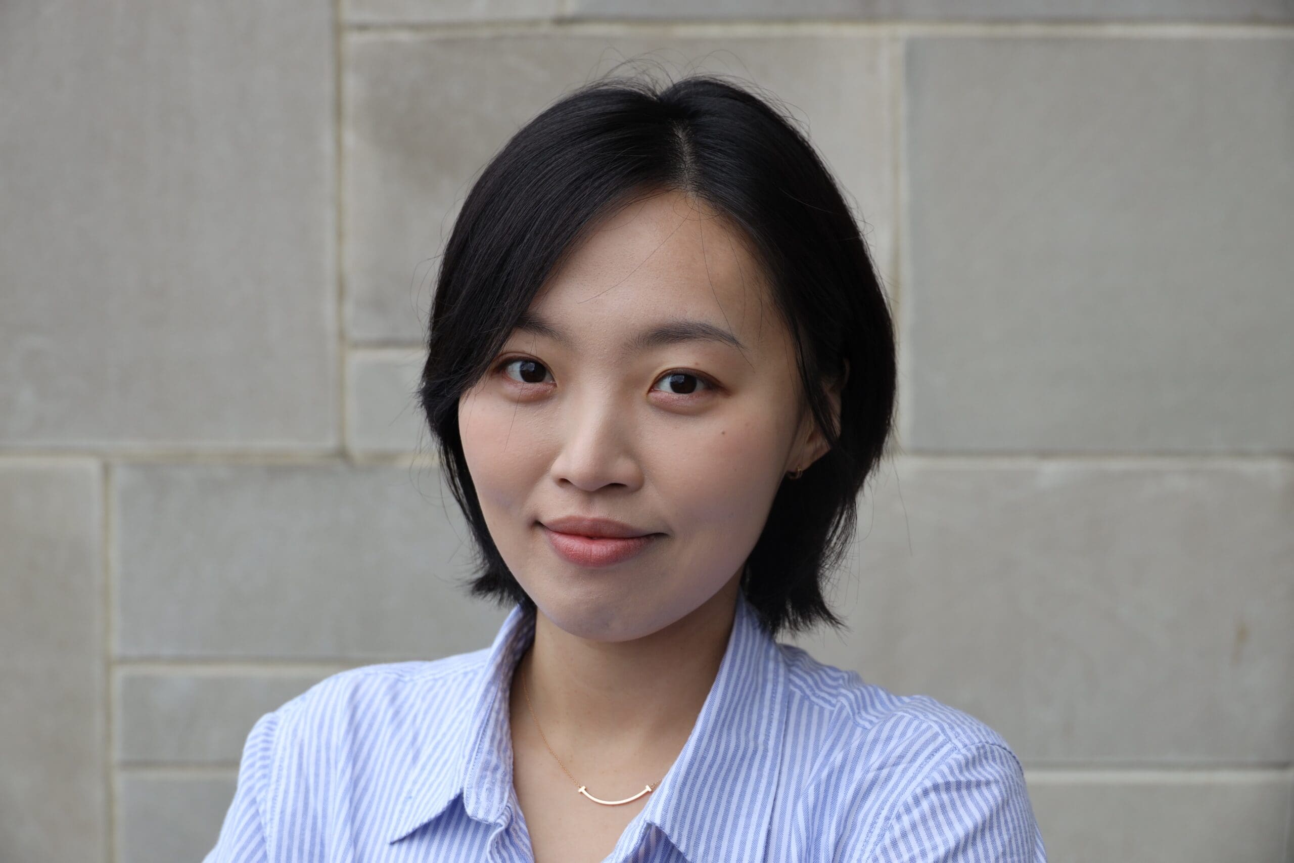 A person with short dark hair wears a light blue striped button-up shirt and a delicate necklace, smiling gently while standing in front of a light gray tiled wall.
