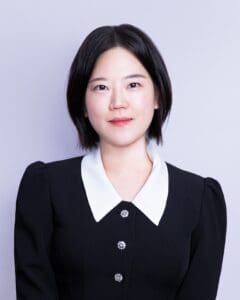 A person with a short dark bob haircut wears a black dress with a white collar and decorative buttons, smiling slightly at the camera against a light gray studio background.
