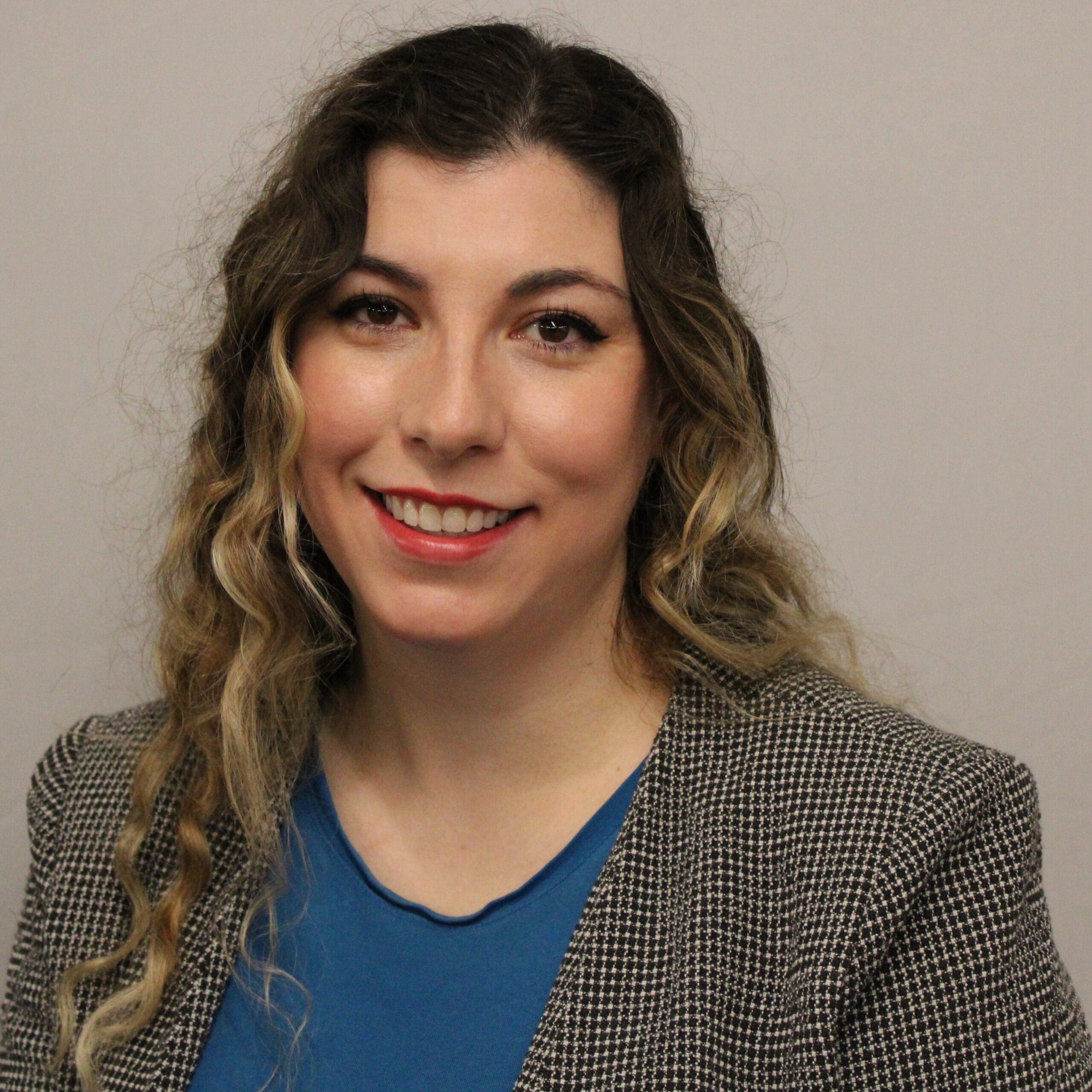 A person with long wavy hair featuring blonde highlights smiles at the camera in a professional portrait. They are wearing a blue top under a patterned blazer against a plain light-colored background.