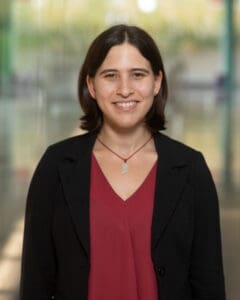 A person wearing a black blazer over a burgundy blouse smiles at the camera. They are standing indoors against a softly blurred background with natural light and a glass-walled corridor.