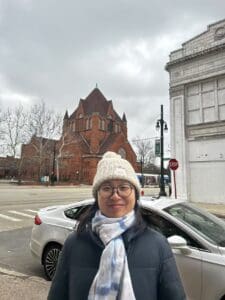 A person wearing a cream knit beanie, glasses, a blue-and-white scarf, and a dark winter jacket smiles at the camera while standing on a city street. Behind them are a red-brick church, a parked silver car, and an overcast sky.