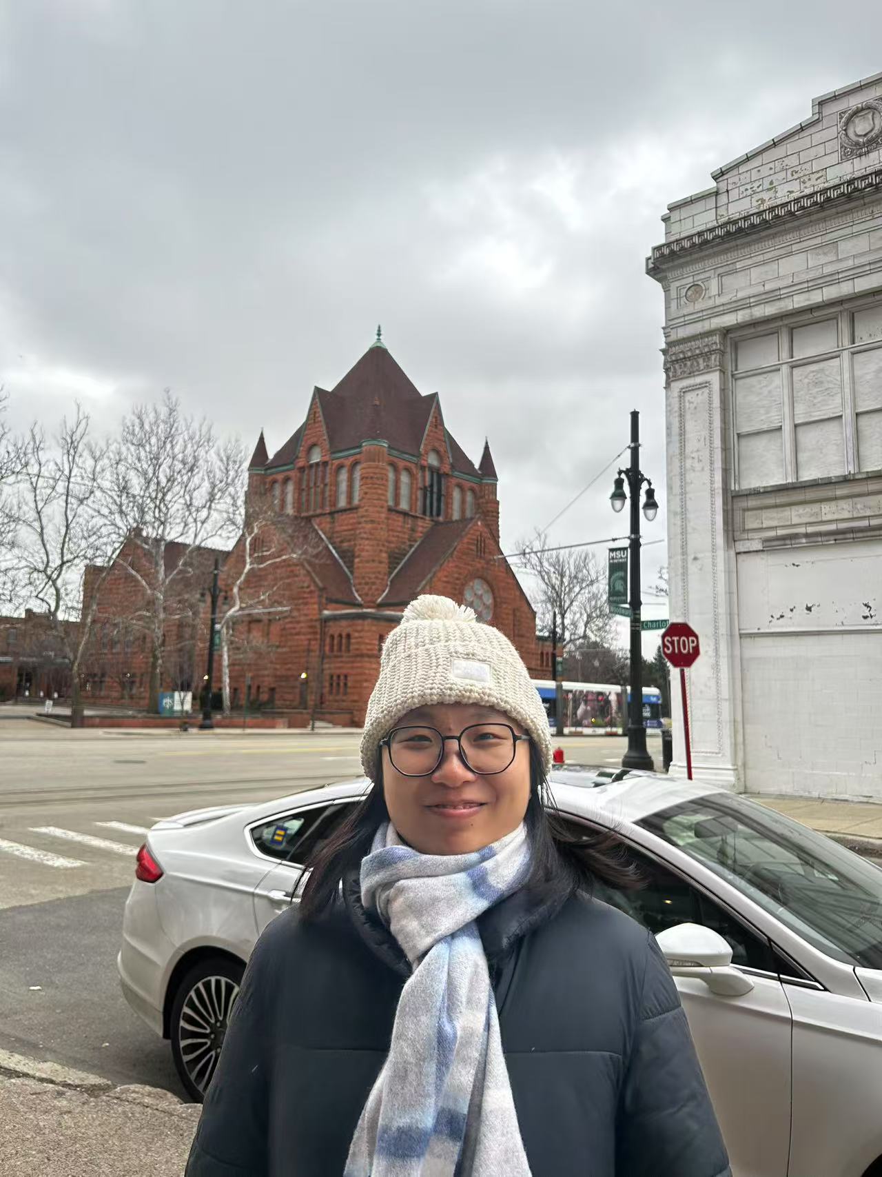 A person wearing a cream knit beanie, glasses, a blue-and-white scarf, and a dark winter jacket smiles at the camera while standing on a city street. Behind them are a red-brick church, a parked silver car, and an overcast sky.