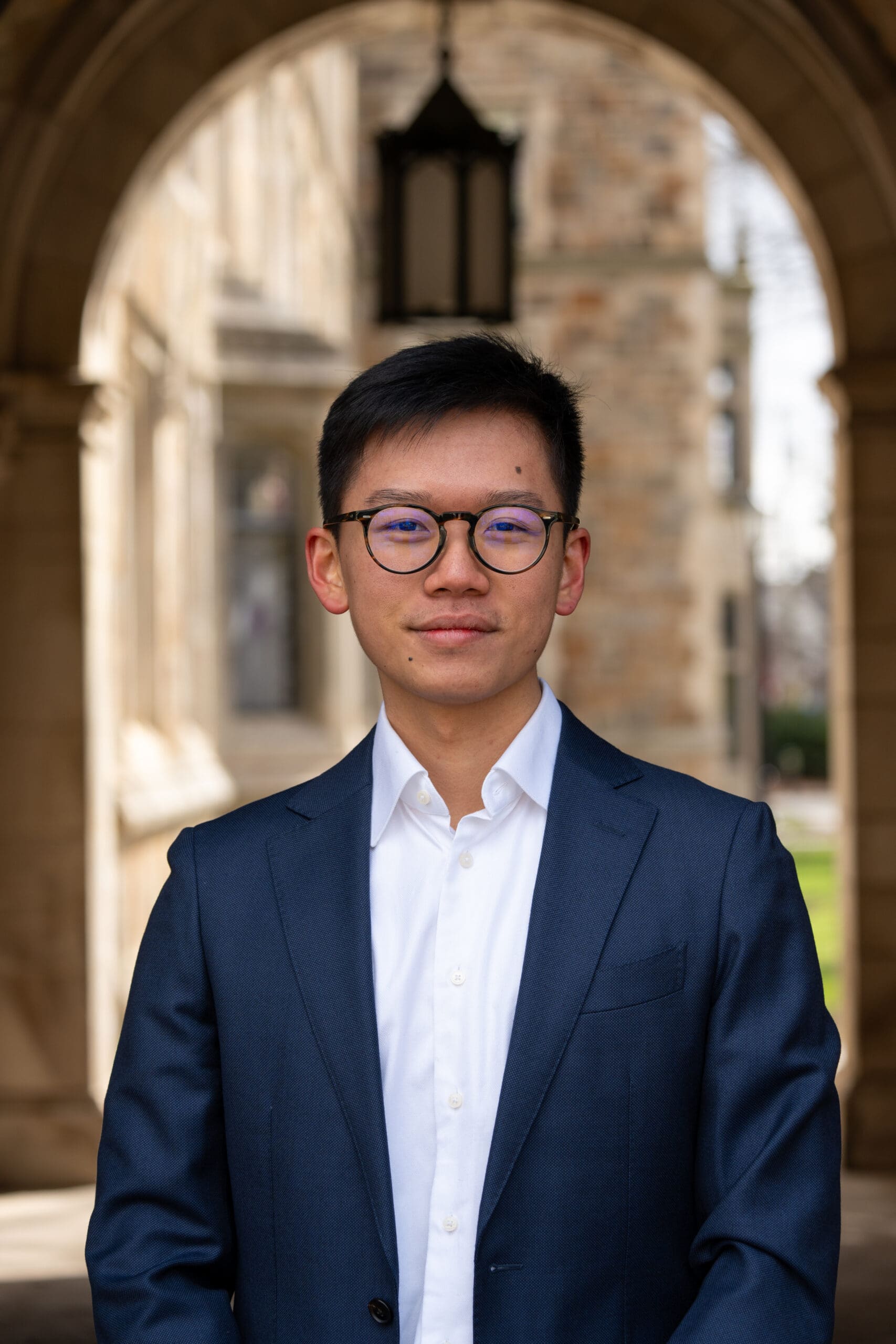 Portrait of a young Asian man in glasses and a navy suit, standing beneath a historic stone archway; calm, confident, professional tone.