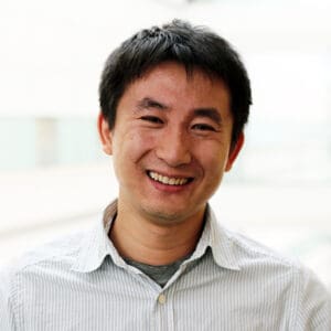 Smiling Asian man with short dark hair in a striped collared shirt, photographed against a bright blurred background; friendly, approachable mood.