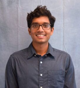 Portrait of a smiling young man with curly dark hair and glasses, wearing a dark button-up shirt against a light gray backdrop.