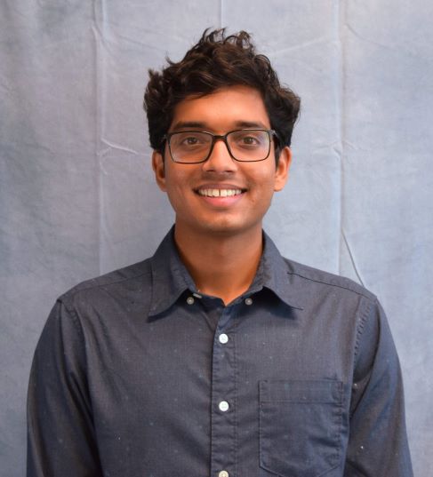Portrait of a smiling young man with curly dark hair and glasses, wearing a dark button-up shirt against a light gray backdrop.